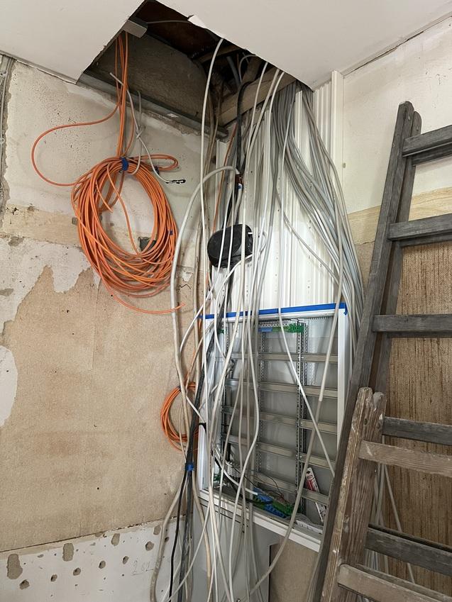 Kabelstrang aus orangefarbenen und weißen Leitungen, die an einer Wand hinter einem Regal und einer Leiter angeordnet sind.