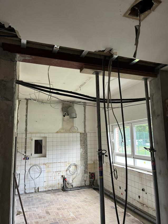 Baustelle mit freiliegenden Rohren und Kabeln, unvollendeter Wandverkleidung und Holzstützen in einem Raum.