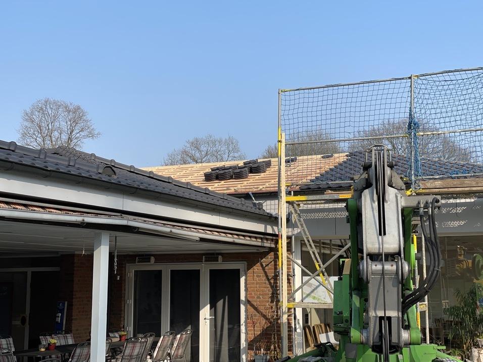 Blick auf ein Dach mit Dachziegeln, auf dem Baugerüst stehen, und einem Baumaschinenarm im Vordergrund.