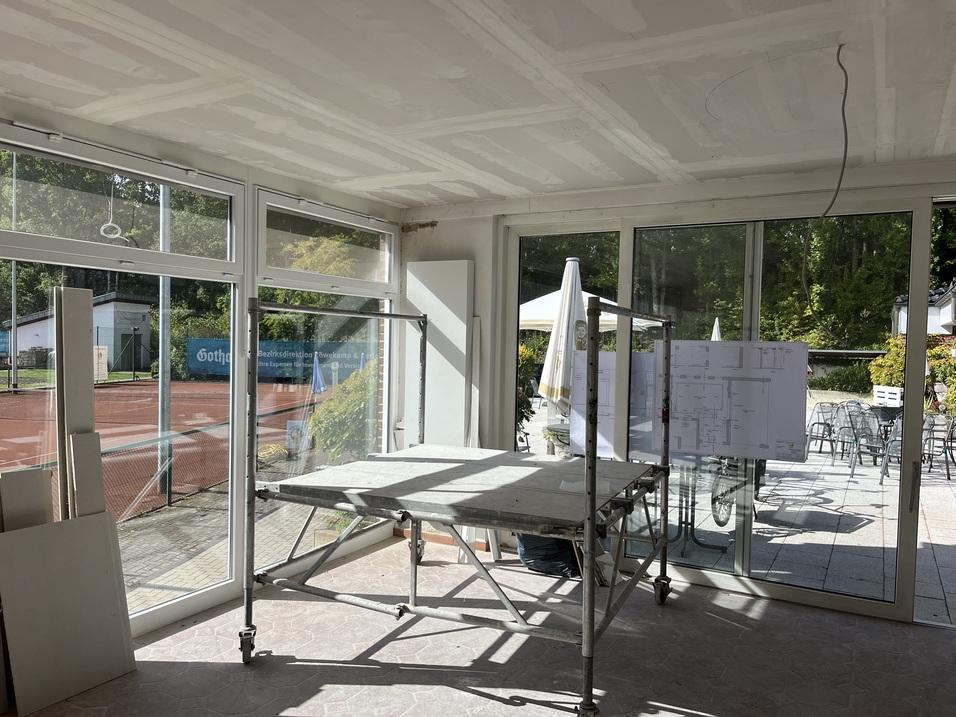 Innenraum mit Baugerüst, großen Fenstern und Bauplänen an der Wand; Außenbereich mit Tennisplatz sichtbar.