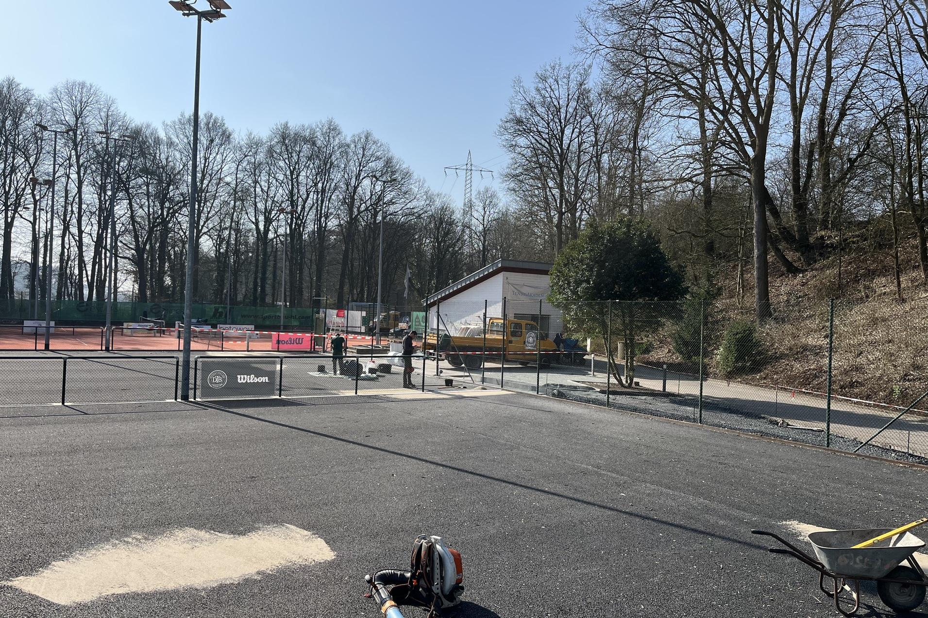 Blick auf einen Tennisplatz mit Bauarbeiten, einer Baumaschine und umgebenden B&auml;umen unter blauem Himmel.