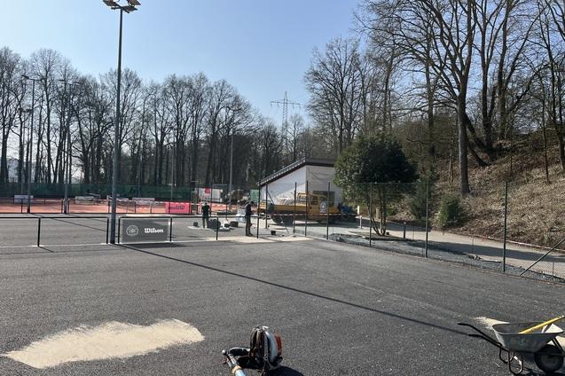 Blick auf einen Tennisplatz mit Bauarbeiten, einer Baumaschine und umgebenden B&auml;umen unter blauem Himmel.