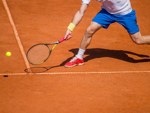 Ein Tennisspieler schl&auml;gt mit einem Racket einen gelben Ball auf einem roten Sandplatz.