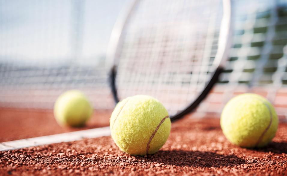 Nahaufnahme von drei Tennisbällen auf einem roten Sandplatz, im Hintergrund ein Tennisschläger und das Netz.