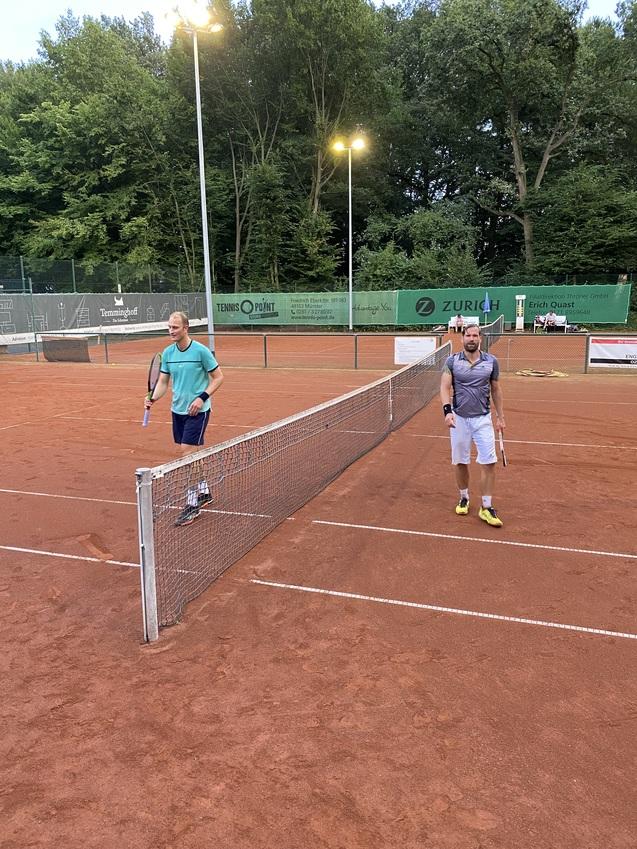 Zwei Tennisspieler stehen an einem Netz auf einem roten Tennisplatz, umgeben von B&auml;umen und Lichtern.