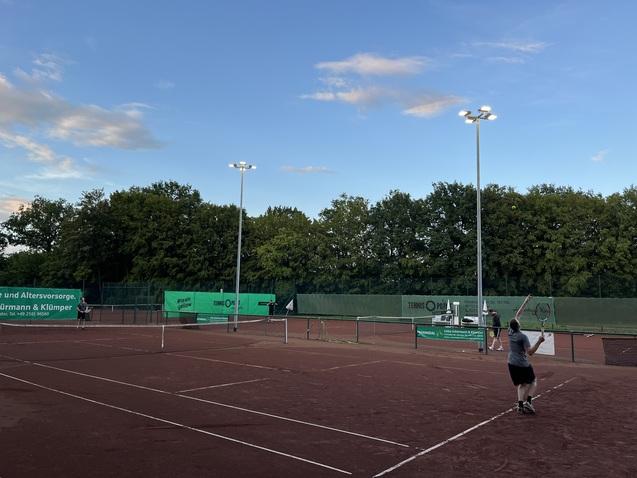 Tennisplatz mit roten Linien, einer Spielerin beim Aufschlag und beleuchteten Pfosten unter einem blauen Himmel.
