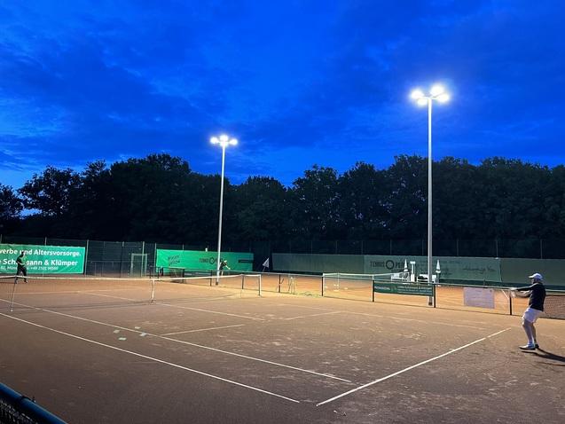 Tennisplatz bei D&auml;mmerung mit Flutlicht und einem Spieler, der sich zur Schlagposition vorbereitet.