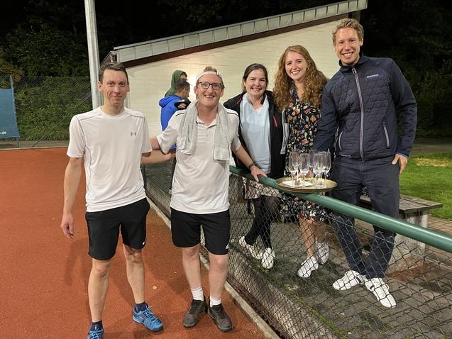 F&uuml;nf Personen stehen auf einem Tennisplatz und halten Gl&auml;ser mit Getr&auml;nken neben einem Tisch mit Snacks.