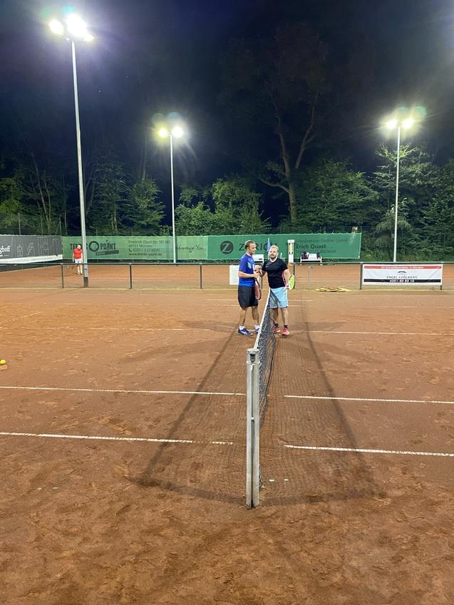 Zwei Personen stehen sich an einem Tennisnetz auf einem beleuchteten Platz gegen&uuml;ber und unterhalten sich.