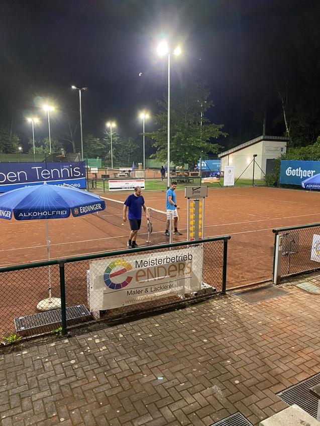 Zwei Personen stehen auf einem Tennisplatz bei Nacht, umgeben von Beleuchtung und Werbebannern.