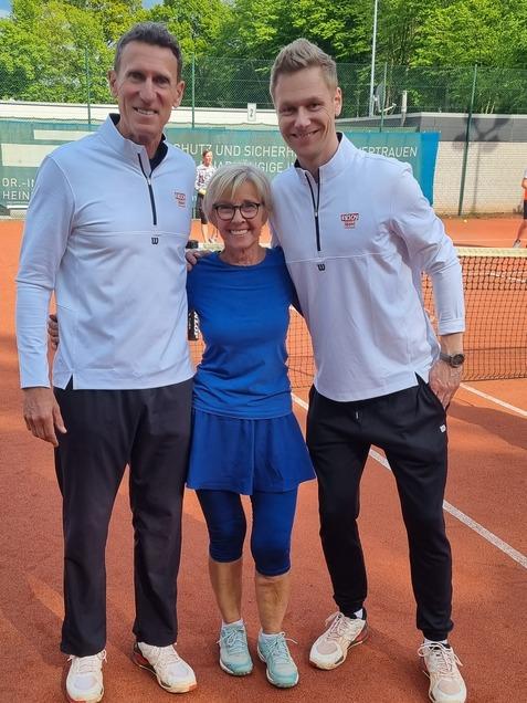 Drei Personen stehen l&auml;chelnd auf einem Tennisplatz, umgeben von gr&uuml;nen B&auml;umen und einem Tennisnetz.