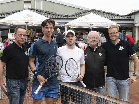 F&uuml;nf M&auml;nner stehen gemeinsam am Tennisplatz, zwei Spieler mit Schl&auml;gern, andere in sportlicher Kleidung.