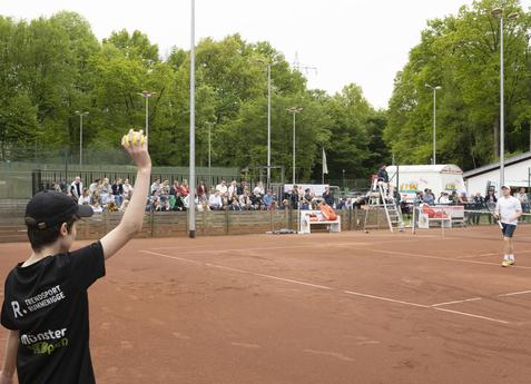 Junge wirft einen Tennisball auf einem Trainingsplatz, im Hintergrund Zuschauer auf B&auml;nken zwischen B&auml;umen.