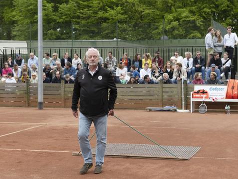 Mann mit grauem Bart zieht eine Besenplatte &uuml;ber einen Tennisplatz, w&auml;hrend Zuschauer im Hintergrund beobachten.