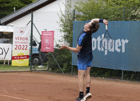 Tennisspieler beim Aufschlag auf einem roten Sandplatz, im Hintergrund Werbebanner und ein Waldgebiet.