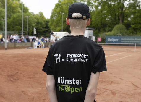 Junger Teilnehmer mit schwarzem T-Shirt steht auf einem Tennisplatz mit Zuschauern im Hintergrund.
