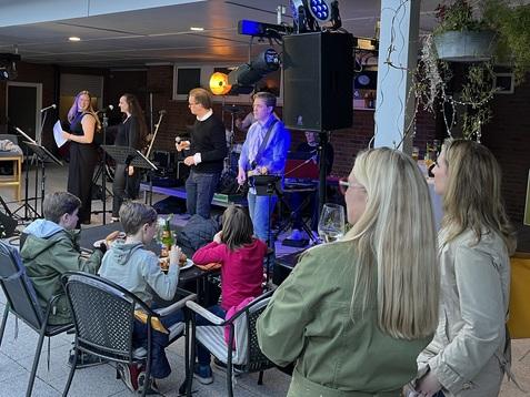 Liveband spielt im Freien, w&auml;hrend Zuschauer in St&uuml;hlen sitzen und Getr&auml;nke genie&szlig;en. Beleuchtung und Pflanzen im Hintergrund.