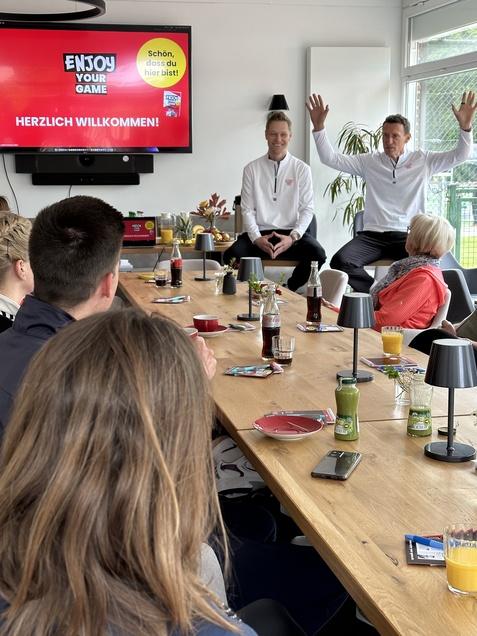 Zwei Personen am Tisch halten eine Pr&auml;sentation vor einer Gruppe, mit einem Bildschirm im Hintergrund, auf dem &bdquo;HERZLICH WILLKOMMEN!&ldquo; steht.