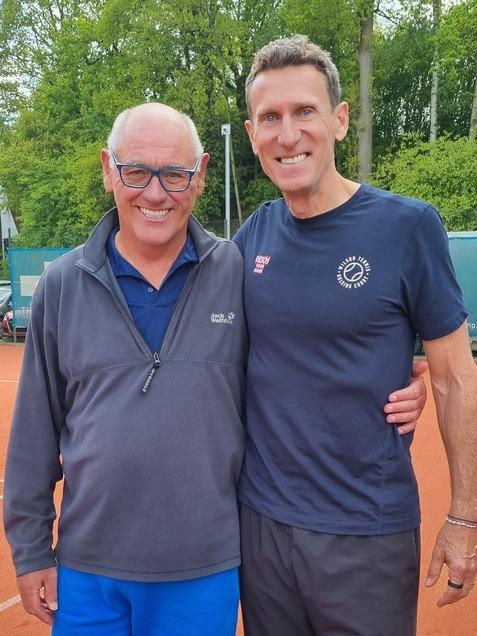 Zwei M&auml;nner stehen auf einem Tennisplatz, einer tr&auml;gt einen grauen Pullover und blauen Shorts, der andere ein schwarzes T-Shirt und schwarze Hose.