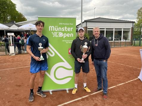 Zwei Tennis-Spieler mit Pokalen stehen neben einem Mann vor einem gr&uuml;nen Banner der M&uuml;nster Open auf einem Sandplatz.