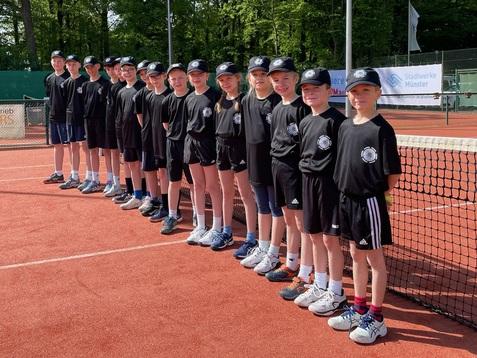 Gruppe von 15 Kindern in Sportkleidung steht auf einem Tennisplatz, umgeben von B&auml;umen und Sonnenschein.