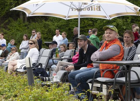 Gruppe von Zuschauern auf St&uuml;hlen unter Sonnenschirmen, w&auml;hrend sie ein Event im Freien beobachten.