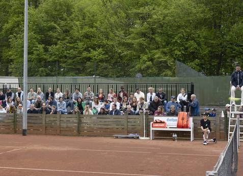 Zuschauer sitzen an einem Tennisplatz, umgeben von B&auml;umen, w&auml;hrend ein Spieler seinen Aufschlag vorbereitet.