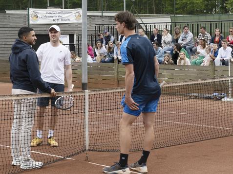 Drei M&auml;nner unterhalten sich am Tennisnetz auf einem Platz, w&auml;hrend Zuschauer im Hintergrund zuschauen.
