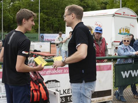 Preisverleihung: Ein Jugendlicher erh&auml;lt eine Auszeichnung von einem Mann auf einem Tennisplatz, Zuschauer im Hintergrund.