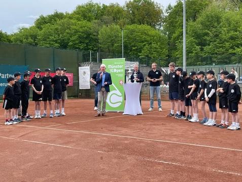 Gruppierung von Kindern in Sportkleidung auf einem Tennisplatz, versammelt um einen Redner und eine Auszeichnung.
