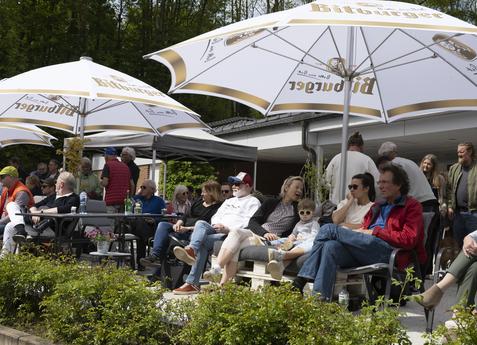 Gruppe von Menschen sitzt unter gro&szlig;en SonnenSchirmen in einem Garten, umgeben von B&uuml;schen und einem Geb&auml;ude im Hintergrund.