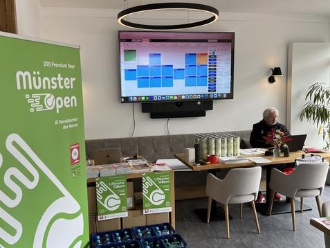 Raum mit Tisch, Monitor und Banner f&uuml;r das M&uuml;nster Open, Person sitzt am Tisch. Getr&auml;nkekisten stehen bereit.