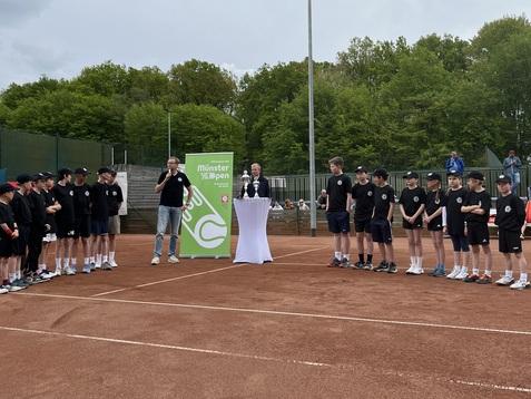Gruppierung von Jugendlichen in Sportkleidung steht um einen Redner vor einem Tisch mit wei&szlig;en Tischdecken auf einem Tennisplatz.
