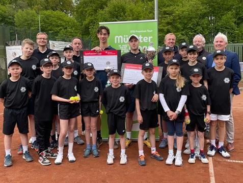 Gruppe von Kindern in schwarzen T-Shirts auf einem Tennisplatz, umgeben von Betreuern und einem Preis.