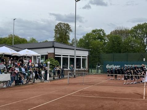 Gruppierung von Sportlerinnen und Sportlern auf einem Tennisplatz mit Zuschauern und einer Restaurantterrasse im Hintergrund.