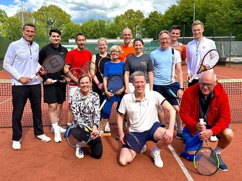 Gruppe von zw&ouml;lf Tennis-Spielern posiert l&auml;chelnd auf einem Platz mit Tennisschl&auml;gern und B&auml;llen, umgeben von B&auml;umen.