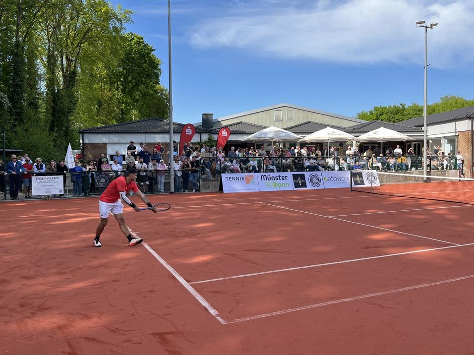 Tennisspieler in rotem Shirt schlägt einen Ball auf einem Sandplatz, im Hintergrund Zuschauer vor einem Vereinsgebäude.