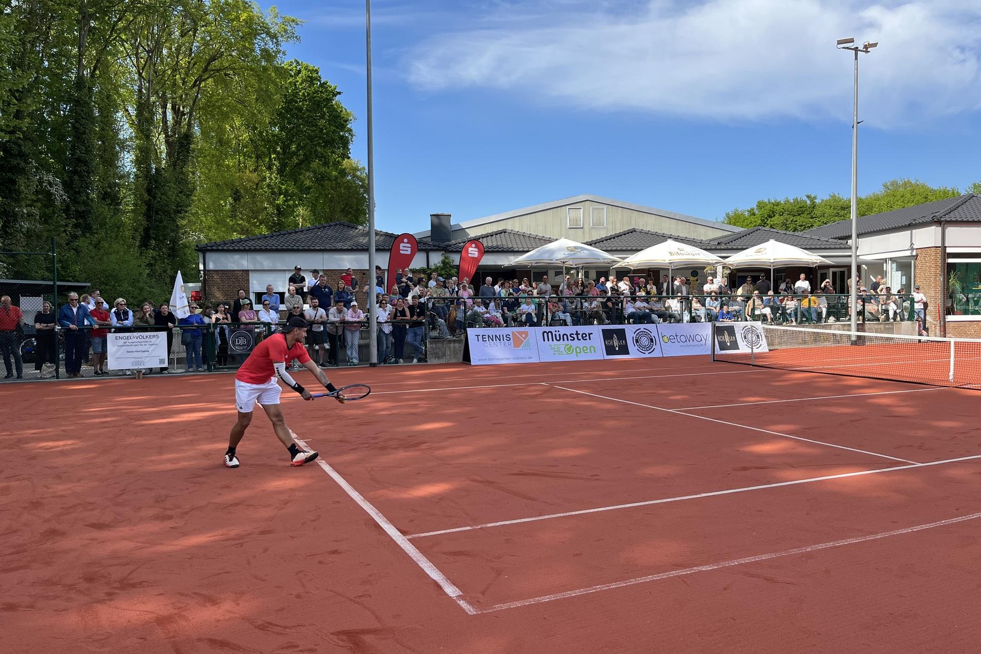 Tennisspieler in rotem Shirt schlägt einen Ball auf einem Sandplatz, im Hintergrund Zuschauer vor einem Vereinsgebäude.