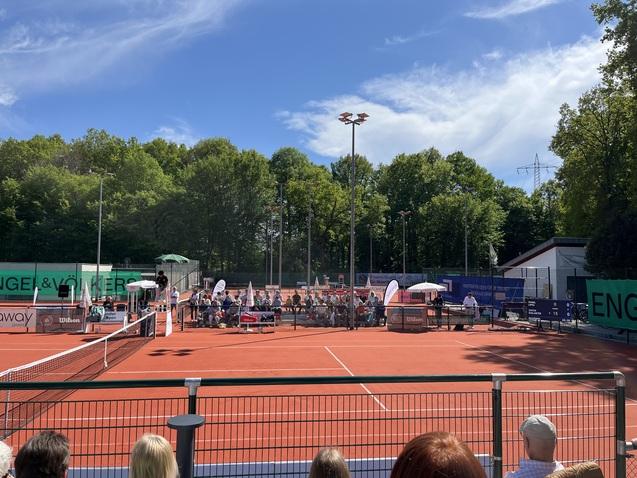 Tennisplatz mit Zuschauern, Schiedsrichter und turniermäßiger Ausstattung unter blauem Himmel und Bäumen im Hintergrund.
