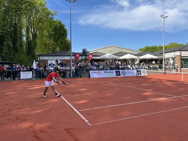 Tennisspieler in rotem Shirt schlägt einen Ball auf einem Sandplatz, im Hintergrund Zuschauer vor einem Vereinsgebäude.