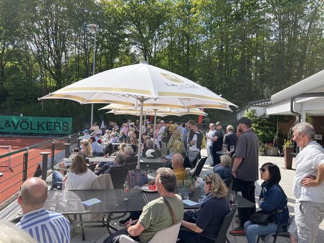 Menschenmenge sitzt und steht unter Sonnenschirmen auf einer Terrasse neben einem Tennisplatz. Grünes Umfeld im Hintergrund.