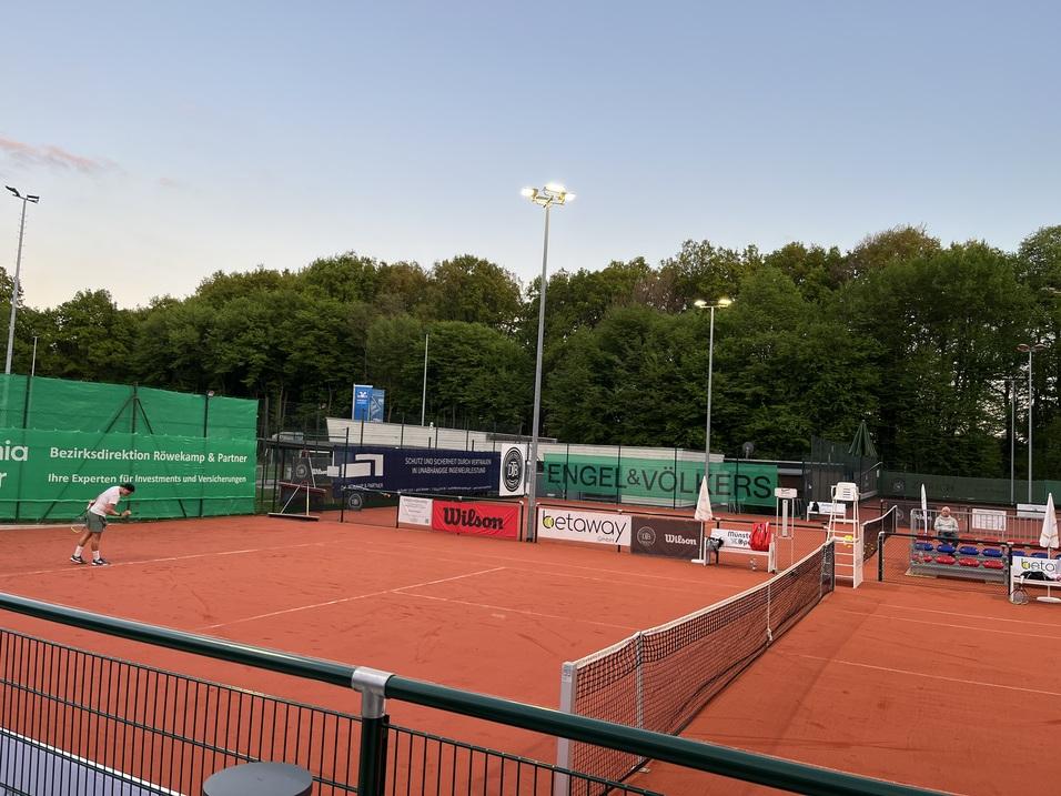 Tennisplatz mit rotem Sand, Werbebanner und einem Spieler in Aktion bei D&auml;mmerung. B&auml;ume und Beleuchtung im Hintergrund.