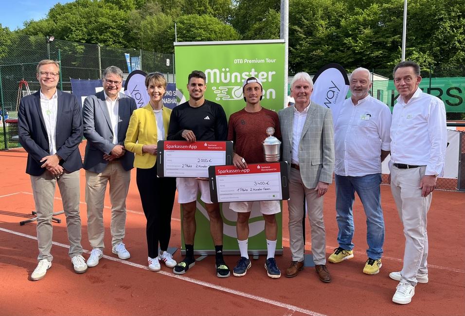 Gruppe von Personen, darunter zwei Tennisspieler mit Siegerchecks und Pokal, vor einem Turnierbanner auf einem Tennisplatz.