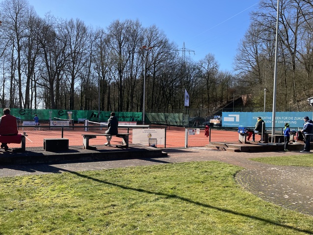 Tennisplatz mit Spielern und Zuschauern, umgeben von B&auml;umen und Wiesen an einem sonnigen Tag.