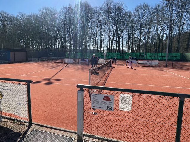 Zwei Spieler auf einem Tennisplatz mit rotem Sand, umgeben von B&auml;umen und Sonnenlicht im Hintergrund.
