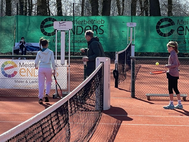 Zwei Kinder stehen auf einem Tennisplatz, einer mit Schl&auml;ger, w&auml;hrend ein Erwachsener Anweisungen gibt.