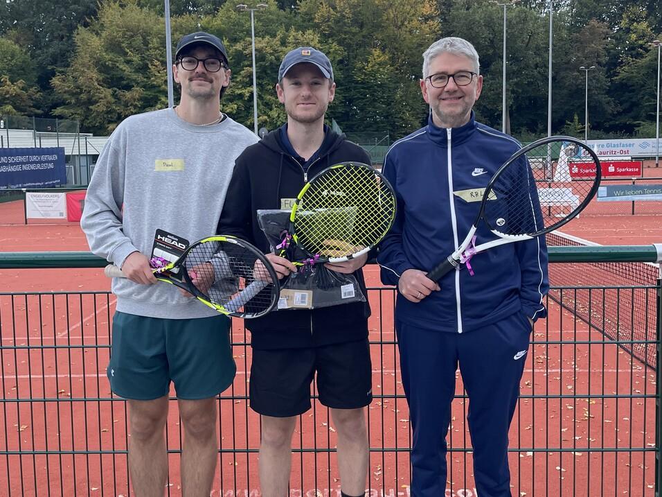 Drei M&auml;nner posieren auf einem Tennisplatz, halten Tennisschl&auml;ger und Medaillen, umgeben von gr&uuml;nen B&auml;umen und Platzlichtern.