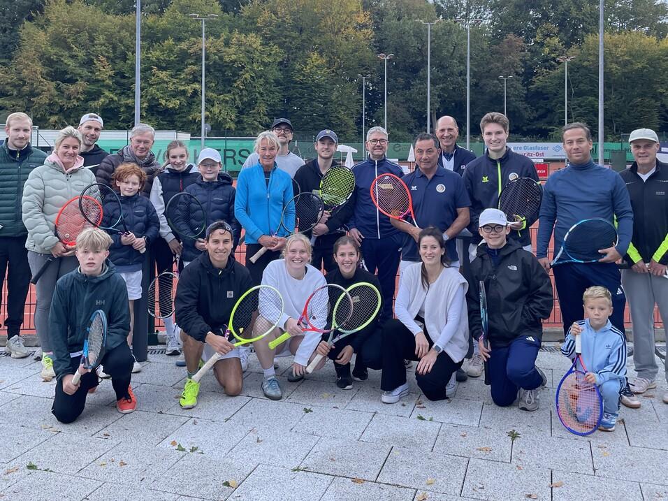 Gruppenshot von 24 Personen auf einem Tennisplatz, alle halten Tennisschl&auml;ger, mit B&auml;umen im Hintergrund.