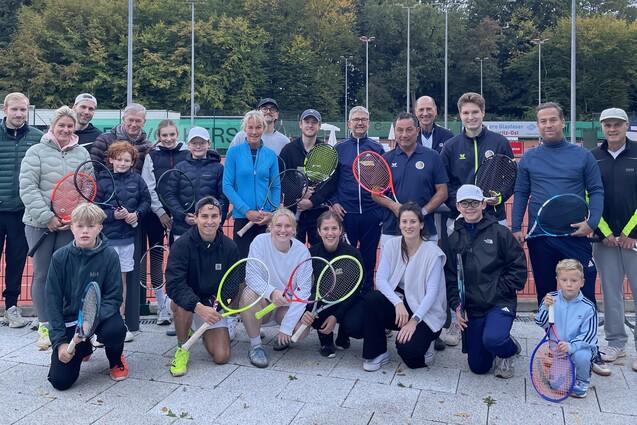 Gruppenshot von 24 Personen auf einem Tennisplatz, alle halten Tennisschl&auml;ger, mit B&auml;umen im Hintergrund.