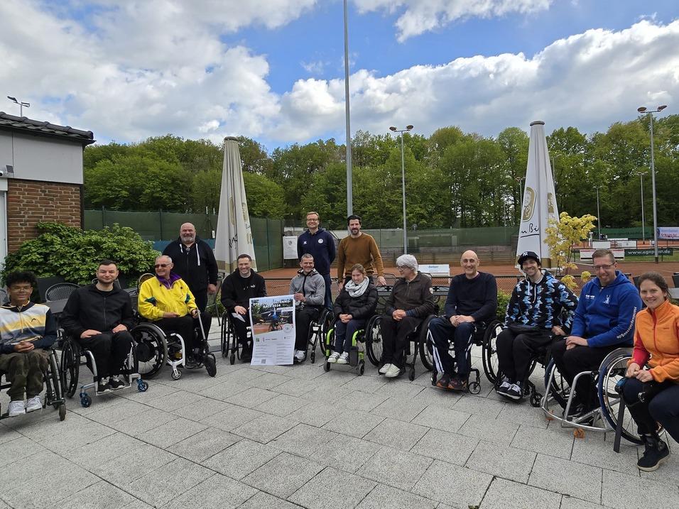 Gruppenaufnahme von 15 Personen im Rollstuhl, die vor einem Sportgelände auf einer Terrasse sitzen.